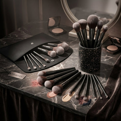 A flat lay on a marble surface of the complete 12-piece Shazory professional makeup brush set, featuring the black terrazzo holder, a travel pouch, and various brushes with powder, liquid, and cream cosmetics, available at getshazory.com