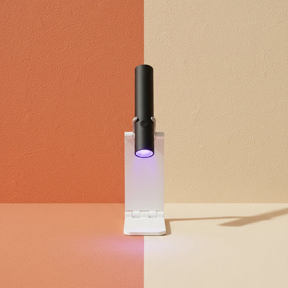 A lifestyle image of the Shazory mini cordless UV/LED nail lamp in black, mounted on its white foldable stand. It is being used to flash cure purple gel nail art on a desk with a passport and accessories, available at getshazory.com