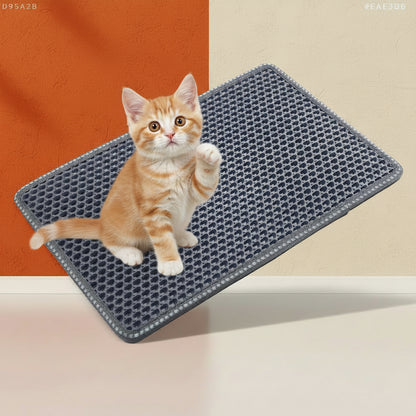 A cute ginger tabby kitten sits on a textured grey Shazory double-layer cat litter trapping mat, which effectively stops litter scatter with its unique MiLi-shape pattern. The mat is displayed against a clean, modern geometric background of muted orange and cream panels. Visit getshazory.com to purchase this essential, easy-clean solution for cleaner pet areas.